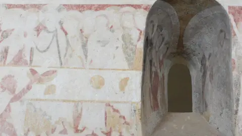 A 12th Century mural of the Last Supper painted onto a wall, with a small window cut into the stonework. The Last Supper is on the left and shows a long table with men with halos sitting behind it, and their legs sticking out beneath. In front of the extreme left is a male figure, in front of the table reaching up to take something off it. The wall paintings continue into the window enclosure and on to the right.