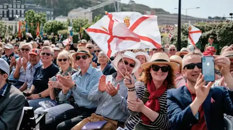 States of Jersey People are sat in a crowd and waving Jersey flags. They are clapping and smiling with a man on the right filming behind where the photo was taken. It is a large crowd with bushes behind with other properties.