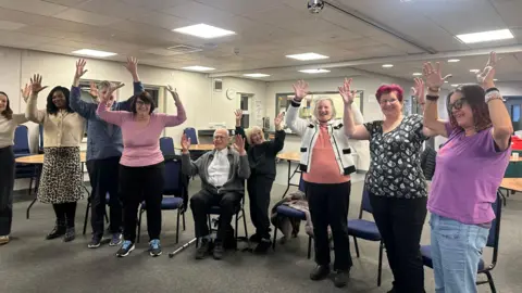 Jessica Bayley/ BBC A group of nine people stood in a line in front of some chairs. All of them have their hands in the air in a jazz hands style.