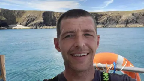 Family Handout A man with short hair standing in a boat with the sea and coastline in the background