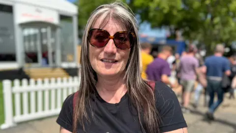 Judith Williamson has long brown and blonde highlighted hair. She is wearing large sunglasses and a black t-shirt, and people are walking behind her outside on a sunny day.