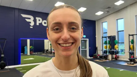The photo shows a woman smiling at the camera. She has brown hair which has been tied back into a ponytail. She's wearing a white top and a silver necklace. She's standing in a gym-like room. There are weights to her right, and a mirror to her left. 