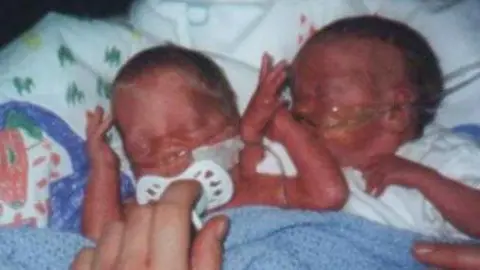 Family handout Two tiny babies are wrapped in a pale blue knitted blanket. Both are snuggled close to each other and have breathing tubes in their noses. The baby on the left has a white dummy in its mouth which is being rested there by an adult hand.