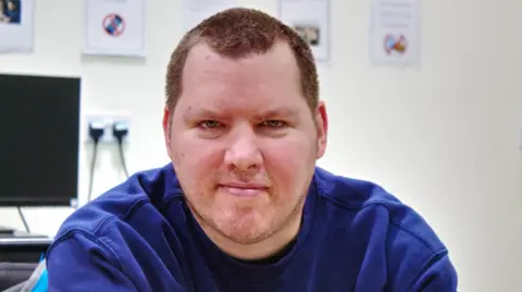 MK SNAP Barry, looking straight at the camera, wearing a blue top, he has short hair, and some stubble on his face. There is a computer behind him to the left. 