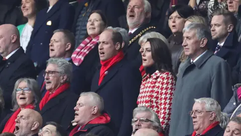 Getty Images Prince William and Catherine singing the national anthems at the Principality Stadium. They are standing in the middle of a crowd in the stands, Prince William wearing a dark suit with a bright red scarf, and Catherine wearing a coat with a red and white houndstooth pattern.