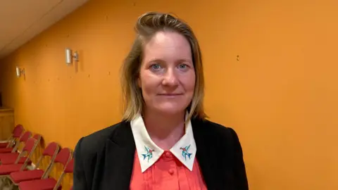 Debbie Beadle looks at the camera. She wears a black jacket with a coral shirt underneath with a white collar - birds are embroidered onto the collar. She stands in front of a yellow wall.