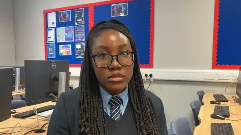 Amy Holmes/BBC Tito has braided black hair and glasses. She wears a black school blazer over a navy jumper, light blue shirt and striped tie. She looks directly at the camera with a neutral expression. She is sitting in a classroom. Behind her are rows of desks with monitors, keyboards, and mice and a display board with red borders featuring various colourful posters.