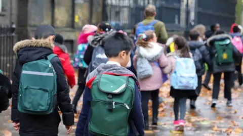 The image shows a group of schoolchildren walking along a pavement covered with leaves, wearing backpacks and coats. They are heading in the same direction towards a school entrance, with an adult walking ahead of them. Tall black railings and brick buildings line the street.