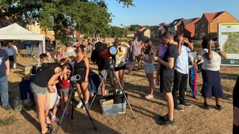 Lyndsay Smith A crowd of people of all ages are gathered together and looking at telescopes in a housing estate, on a green.