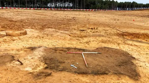 Oxford Cotswold Archaeology A large archaeological excavation site of sandy coloured soil extending out to pine trees in the distance. In the foreground is a much darker rectangle of soil, the remains of a funeral pyre. Resting in its middle are two red and white coloured measuring sticks. 