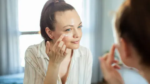 Woman with dark hair applies a facial skincare product in the mirror.