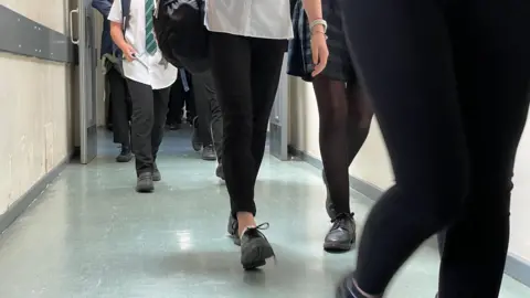 Children walking in a school corridor. Their feet and legs are visible and they are walking in a group. Some are wearing trousers and a girl is wearing black tights with a skirt. They all have black shoes.