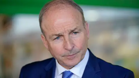 PA Micheal Martin wearing white shirt, blue tie and blue jacket stares ahead waiting to answer questions from the media