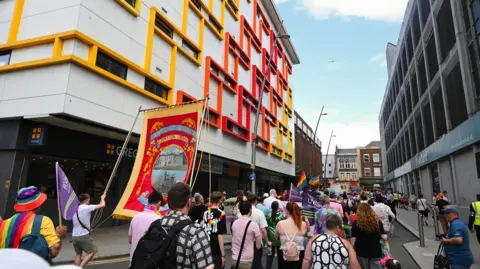 Haydn Brown A crowd of people marching away from the camera. They are walking down a street lined with large buildings, the one on the left is mostly white with colourful yellow, orange and red lines around the windows. There are Pride flags being held up, as well as purple and green Unison flags. A small group of people are holding up a large red and yellow union banner which reads 'National Union of Mineworkers' with a painting in the middle.
