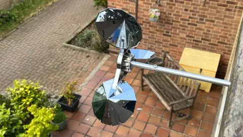 Darren Rozier/BBC A shiny metallic bird-scarer device placed above an outdoor space, including a garden bench and patio area.