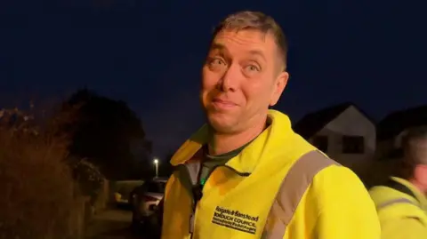 A man in high-vis looks at camera whilst another man, similarly dressed, pulls a bin along behind him.
