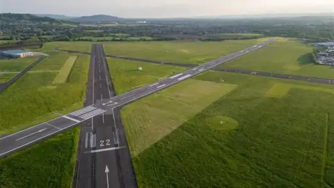 Gloucestershire Airport Aerial view of Gloucestershire Airport's two intersecting runways, flanked by grass with rolling views visable in the background.