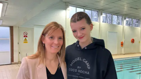 A woman with straight blonde hair and a pick blazer is smiling at the camera and standing next to a teenage girl who has her hair pulled back and is wearing a blue hoody. They are both standing next to a swimming pool