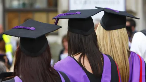Three female university graduates stand with their backs to the camera. They're wearing the traditional graduation gown and hat.