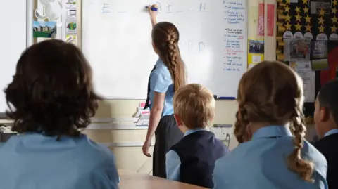 Getty Images Students in a classroom, pictured from behind. They wear blue shirts. One is standing at the whiteboard writing.