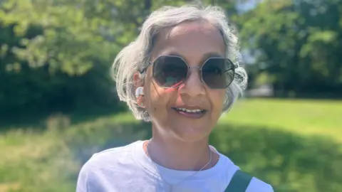 BBC / Elizabeth Baines A woman with white hair smiles at the camera. She is wearing sunglasses, earphones and a white top. Behind her, green space is blurred.