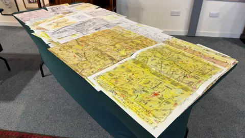 A table covered by pieces of paper with a variety of images in them. Many of the works have a yellow background with images drawn on top.