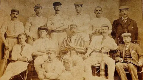 Selkirk Cricket Club an old image of a team of cricketers all posing as a team. the picture is a washed out yellow colour. the main in the middle of the team is wearing a striped blazer. 