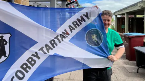 A woman holds out a blue flag that reads Corby Tartan Army in a pub garden. She is smiling at the camera. She wears a green T-shirt, dark trousers and has short brown hair. 