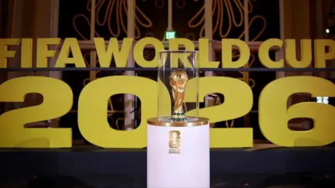 The FIFA World Cup trophy inside a case on a podium infront of a sign naming the 2026 tournament.
