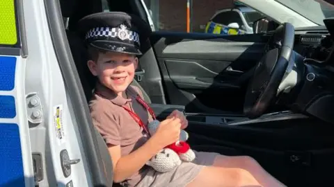 Eli, a seven-year-old boy is sat in the driver's seat of a police car, wearing a police hat. 