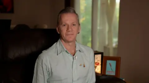 Keir MacKenzie / BBC A man wearing a light blue shirt stood in his living room. Photo frames with a book camera sit in the background.