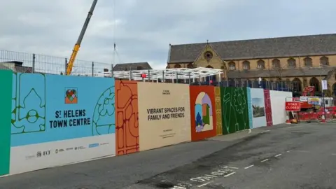Multi-coloured boarding on the side of a construction site which reads: "St Helens town centre" and "vibrant spaces for family and friends". A church building is in the background with fencing and a red sign in front which says "Road closed" in white writing.