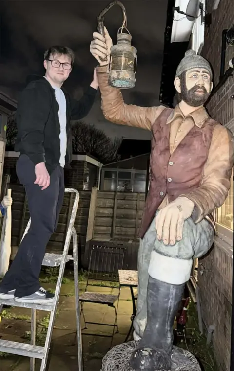 Kyle Wilcock Kyle Wilcock standing on a steel step-ladder in the dark at the back of his house. He has glasses and is wearing dark trousers, a grey tshirt and a black jacket and his left hand is touching the right arm of the statue which holds up a lamp. The statue is of a miner with a beard, grey woollen hat, brown shirt, dark red waistcoat, grey trousers and wellies. Its left foot it up on a log or barrel and its left hand rests on its knee