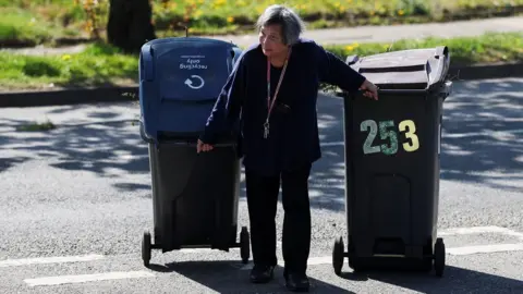 A woman pulls her wheeled bins to a Mobile Household Waste Centre where residents can dispose of their rubbish. She is wearing a dark top and trousers and has a lanyard around her neck. She is pulling two bins and stooping slightly. The sun is out and there is grass behind her and a tree trunk.