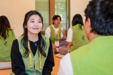 BBC / Hosu Lee A participant wearing a light green vest and yellow lanyard sits on a floor cushion in a temple hall, facing another participant. Several others are visible in the background