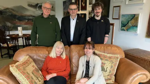 The mothers are sitting on a leather sofa with the father's behind them accompanies by Orson Tindal-Draper, one of Callum's brothers. They are all smiling.
