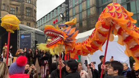 Men hold a lion puppet on long red poles as they walk towards a stage. They are surrounded by people filming it on their phones