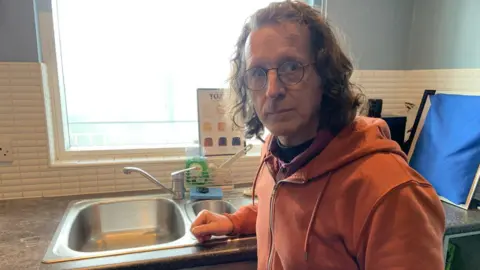 A man with shoulder length brown hair is wearing glasses and an orange hoodie. He's standing in a kitchen by a sink