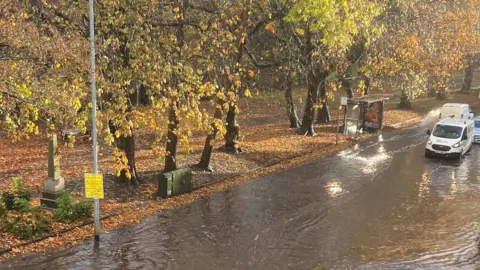 LDRS A flooded road, lined with trees. There is a white van driving into the flood water in the right hand side of the image and another white van leaving the flooded part of the road behind it.