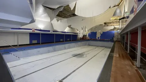 Pughs Auctions An empty swimming pool in a closed centre. The roof lining is falling down towards the pool. The lights are on, but the room is empty. 