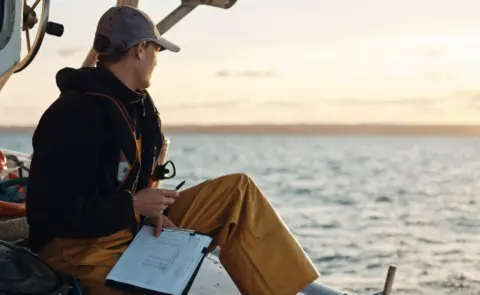 Matt Sharp Freddie sat on the edge of a boat with a clip board and pen