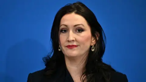 Getty Images Emma Little-Pengelly smiling. She is wearing red lipstick, a black jacket and gold earrings. She has long black hair. The background behind her is blue.