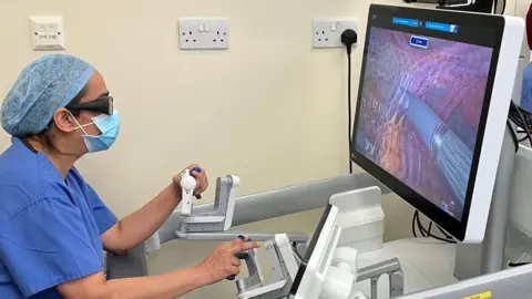 A female consultant in blue scrubs, glasses and face mask operates a machine which carries out robotic hysterectomies.