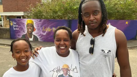 Irene stands between her son Elliot and a younger child. She and the younger child are wearing white t-shirts with a picture of Eddie on them and his name. The three are holding each other and smiling brightly in front of the mural. 