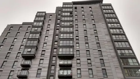 LDRS A photo of the grey and black tower block taken from the ground next to it, with the tower looming over the photographer against a white cloudy sky.