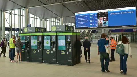 Pacemaker Passengers walking about Grand Central Station in Belfast. In the foreground some ticket machines. In the background a large screen with departure times on one side and "Welcome to Grand Central" on the other.