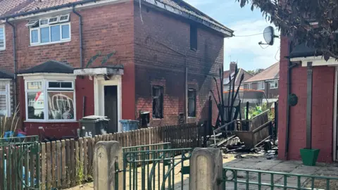 BBC / Michael Nagasaka The photo shows a brick building and its drive. There is visible fire damage to the side of the building, which is covered in black soot. A tree has also been scorched. 