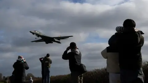Six people can be seen in the foreground taking photographs, as a USAF B-1 bomber approaches to land at RAF Fairford airbase. The photographers all wear coats, with all bar one in hats as well.