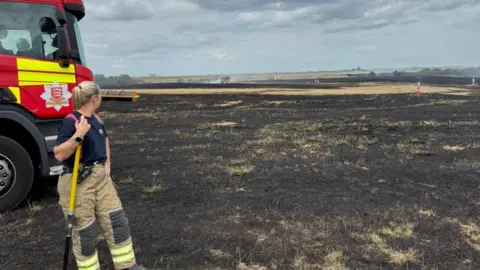 Essex County Fire and Rescue Service A firefighter stands in a burnt field holding a shovel, with a fire engine nearby. The sky is partly cloudy, and the landscape in the background shows more burnt areas.