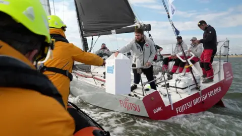 Shaun Whitmore/BBC The British Soldier boat at sea can be seen from behind. The crew are sitting aboard. One is holding a tall white box which he reaches over the railings with. He is handing it over to a man in a yellow jacket on an orange lifeboat. 